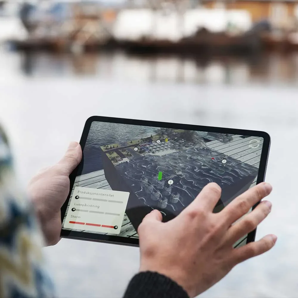 An image of the hands of a person holding an ipad. The person is standing outside by the ocean, and the screen on the ipad shows a 3D model of a coast line hosting three fish farming methods. The methods have a visual meter showing the impact of three parameters: production intensity, salmon lice, storm.